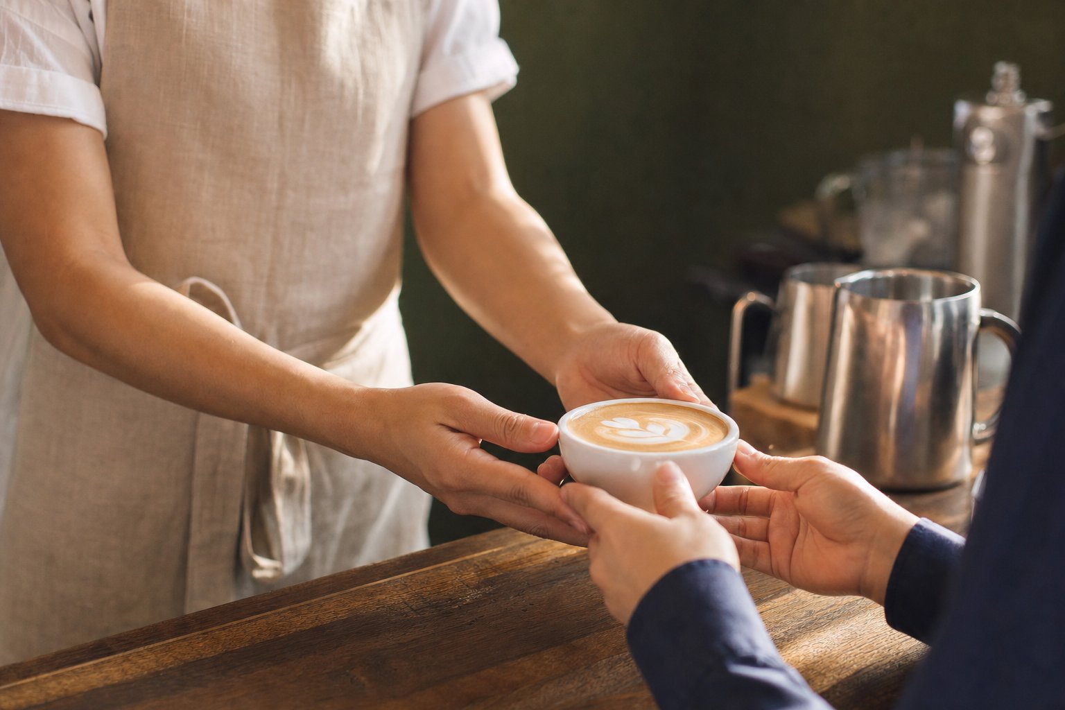 A barista in a linen apron gently hands a flat white across a wooden café counter